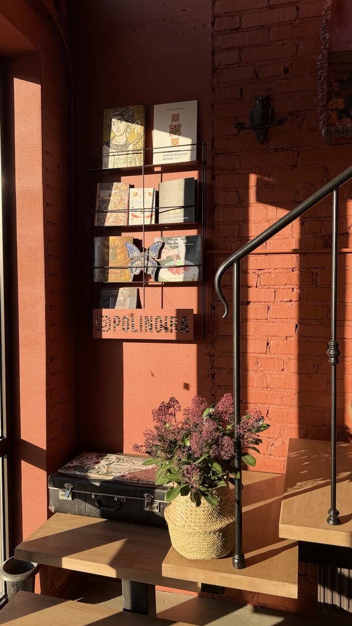 Inside the POLINO studio — a shelf of mosaic art books above a vintage suitcase and dried blooms, lit by low sun on a terracotta brick wall